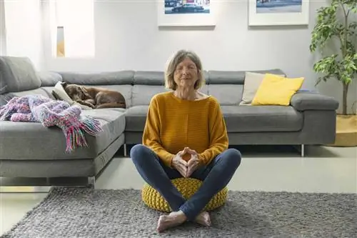 Пожилая женщина сидит на желтой подушке с закрытыми глазами и медитирует.