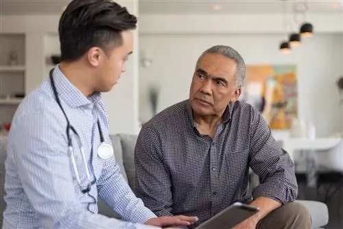 Врач азиатского происхождения и пожилой пациент находятся в помещении дома мужчины. Врач разговаривает со своим пациентом о его депрессии и беспокойстве, показывая ему планшет.