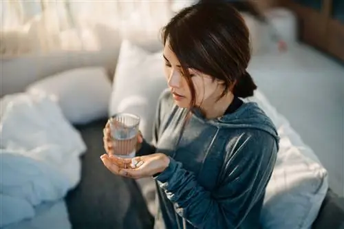 Молодая азиатка сидит на кровати и чувствует себя больной, принимая лекарства в руке со стаканом воды - стоковое фото