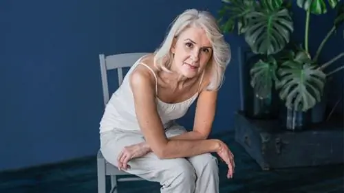 женщина сидит в кресле и смотрит в камеру, растение позади нее